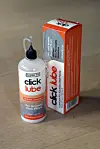 Flaske og eske med Click Lube smøremiddel plassert på et parkettgulv.