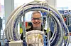 Person med vernebriller smiler bak en stor kveil med malerslanger og sprøyteutstyr i et verksted.