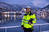 Person i gul vernejakke og hjelm står ved vannet med Tromsø og snødekte fjell i bakgrunnen.