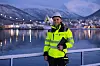 Person i gul vernejakke og hjelm står ved vannet med Tromsø og snødekte fjell i bakgrunnen.