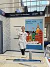En maler i hvitt arbeidstøy står foran en fargerik byillustrasjon ved en arbeidsbenk.